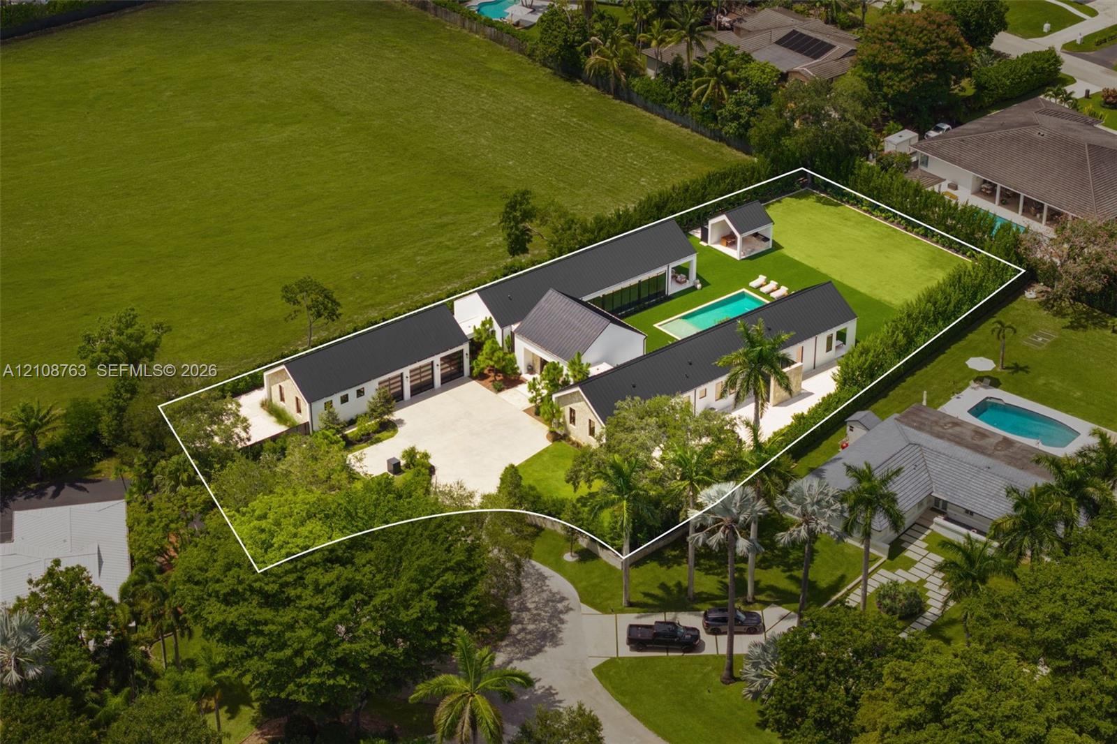Estate featuring a multi-structure layout with metal roofing, a swimming pool, detached gazebo, expansive paved motor court, and manicured grounds enclosed by privacy hedges