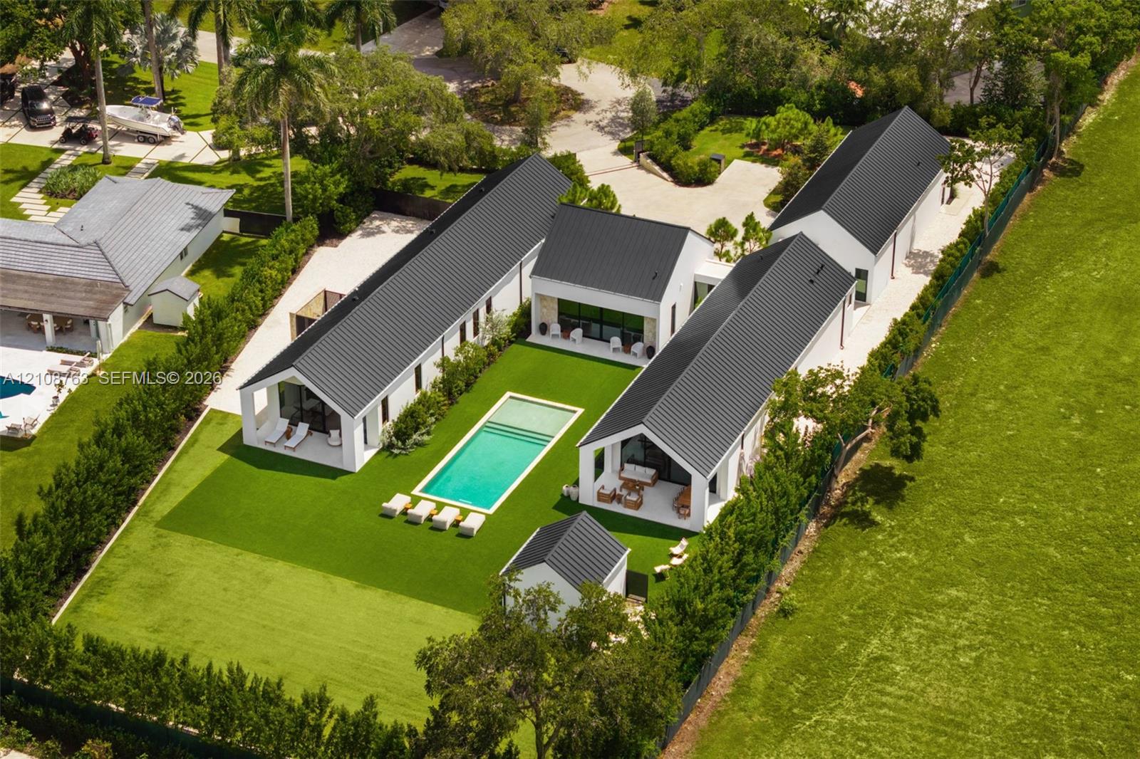 Modern estate featuring multiple gable-roofed structures, a central rectangular swimming pool, concrete paved motor court, artificial turf lawn, and perimeter privacy hedging