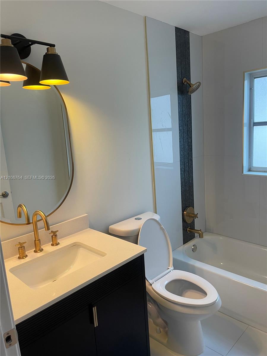 Bathroom featuring a dark wood-finish vanity with a white countertop, oval wall-mounted mirror, brass-toned fixtures, and a tub with a tiled shower surround accented by a decorative dark vertical inlay Bathroom featuring a dark wood-finish vanity with a white countertop, oval wall-mounted mirror, brass-toned fixtures, and a tub with a tiled shower surround accented by a decorative dark vertical inlay