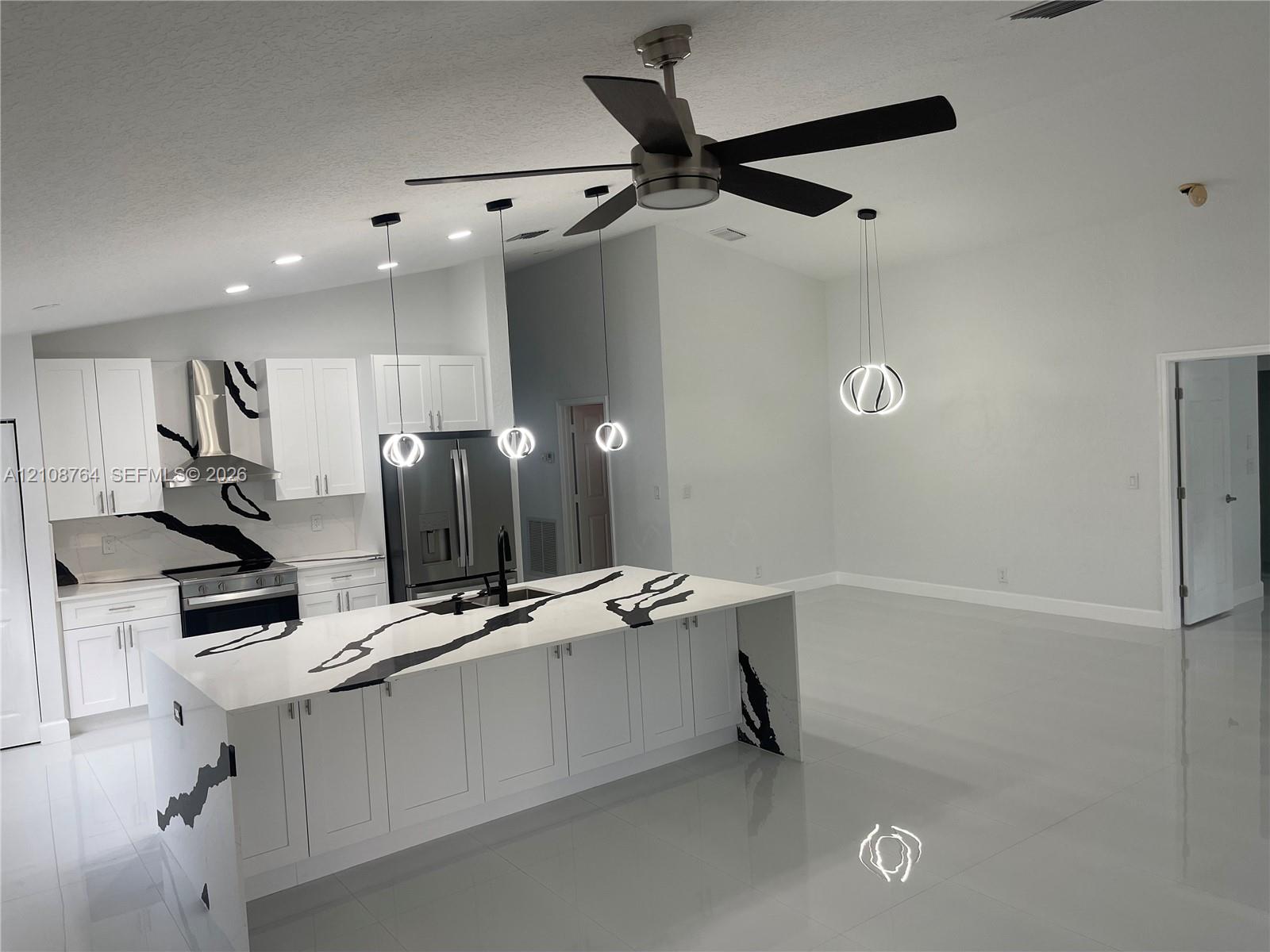Kitchen featuring a central island with a waterfall countertop, white shaker-style cabinetry, a stainless steel range hood, recessed ceiling lighting, and suspended globe-style pendant fixtures Kitchen featuring a central island with a waterfall countertop, white shaker-style cabinetry, a stainless steel range hood, recessed ceiling lighting, and suspended globe-style pendant fixtures