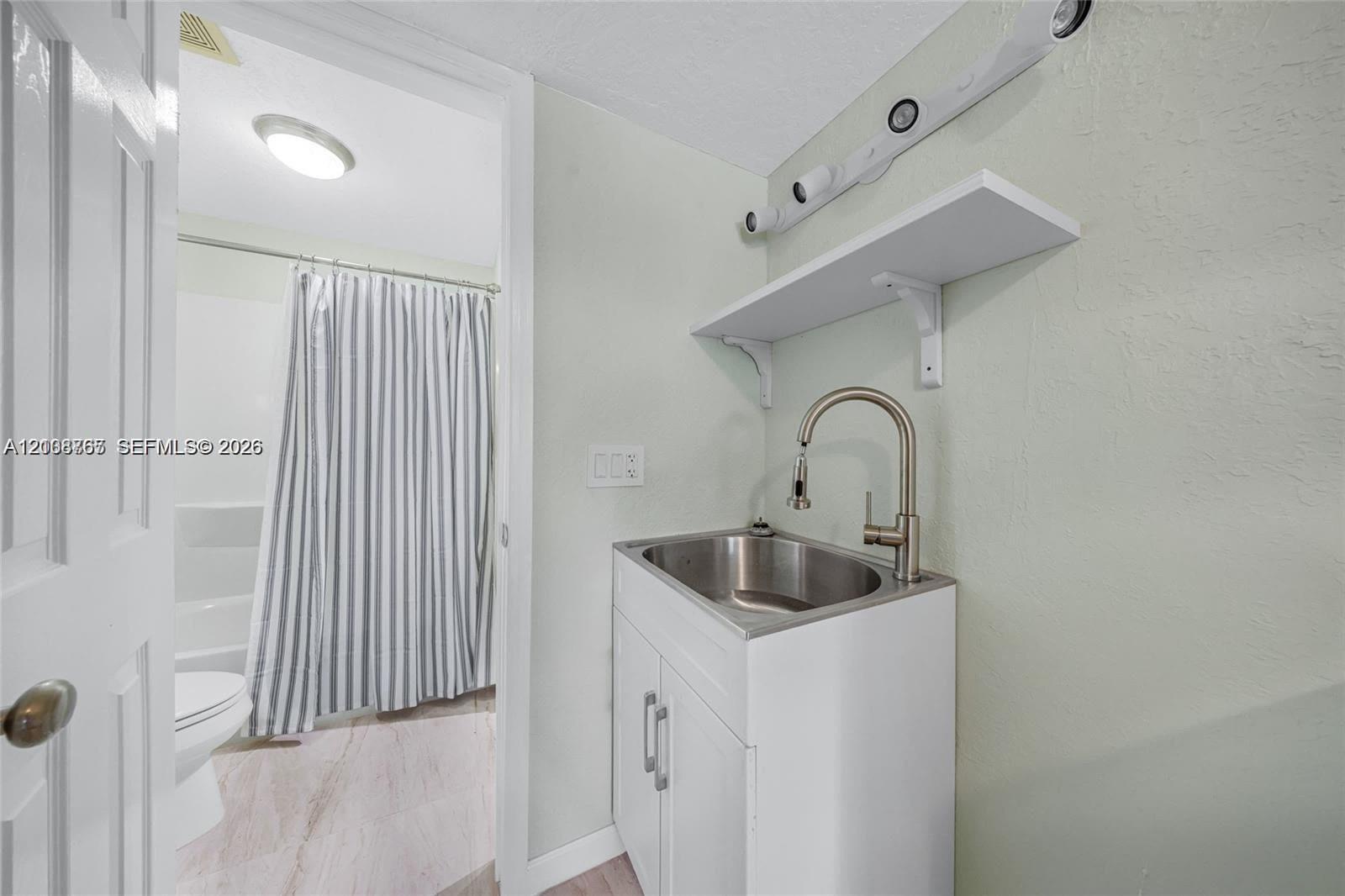White vanity featuring a stainless steel drop-in sink, high-arc gooseneck faucet, and wall-mounted shelving, paired with a multi-light fixture