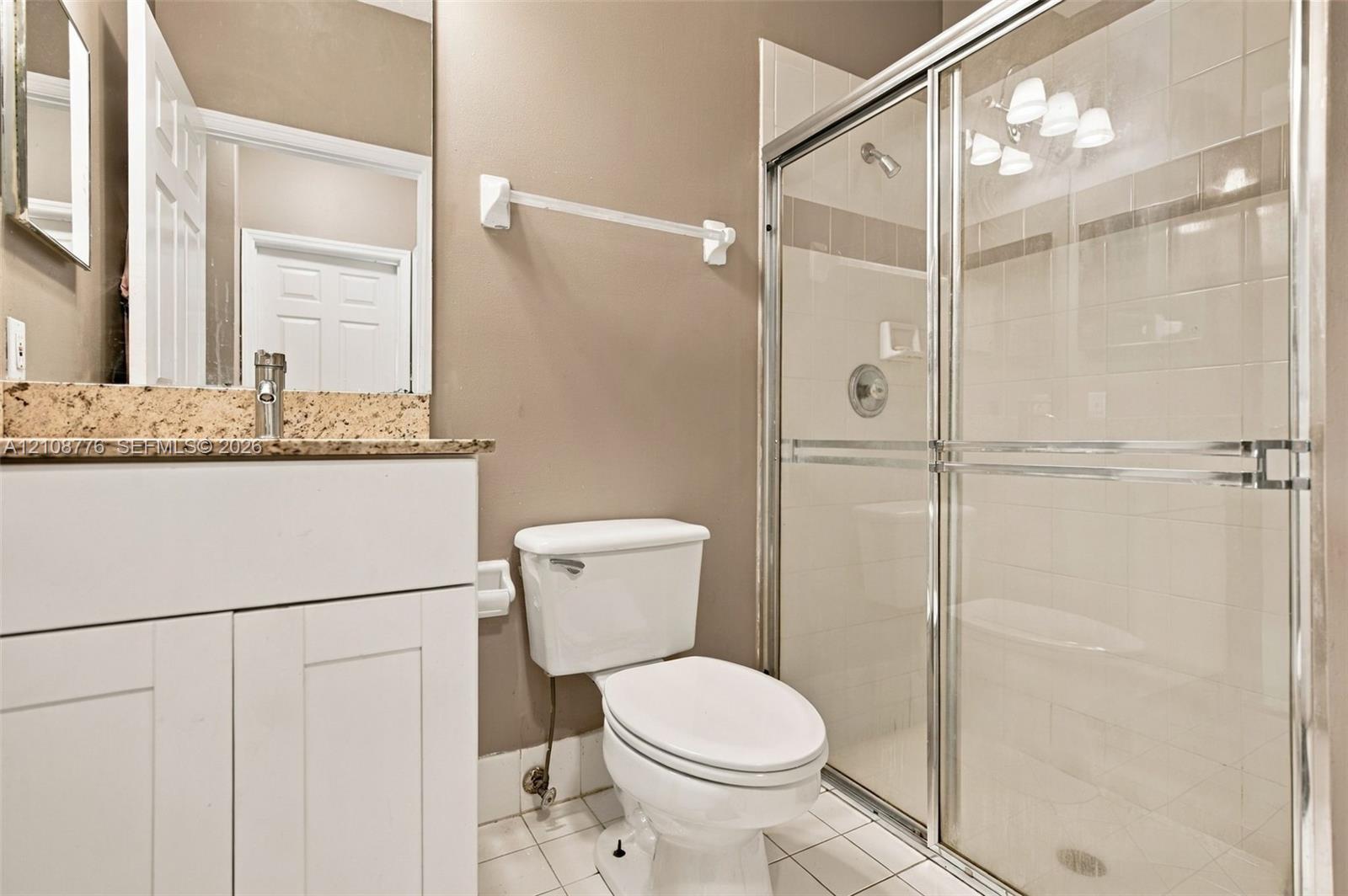 Bathroom featuring a white vanity cabinet with a speckled stone countertop, a glass-enclosed shower with tiled surround, and ceramic tile flooring