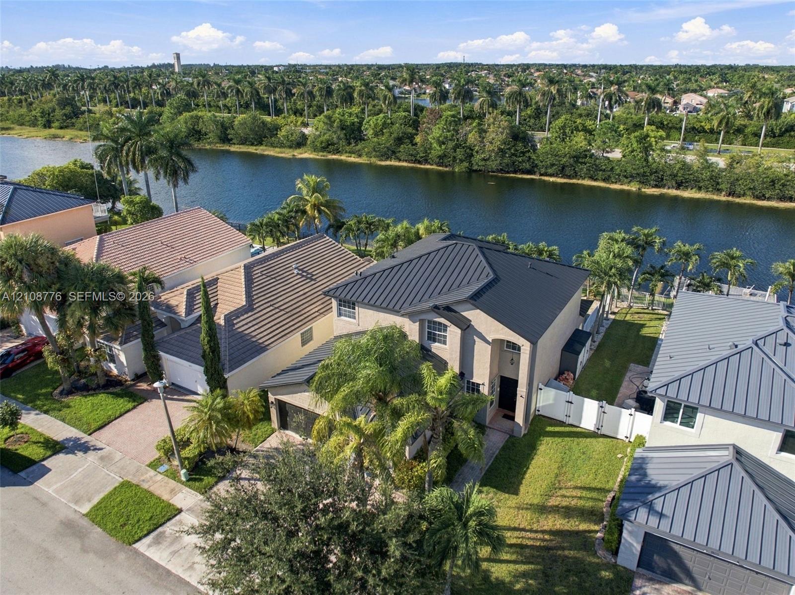 Stucco exterior residence featuring a metal roof, paved driveway, white vinyl perimeter fencing, and water frontage