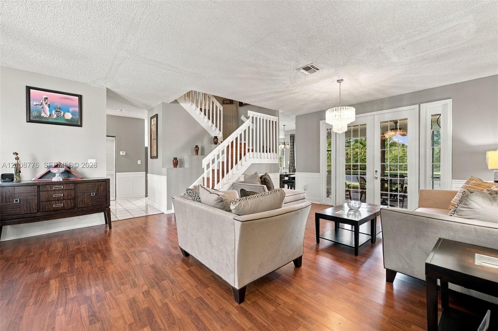 Living area featuring wood-finish flooring, a white staircase with a wooden handrail, glass-paned double doors, wainscoting, and a multi-tiered crystal chandelier
