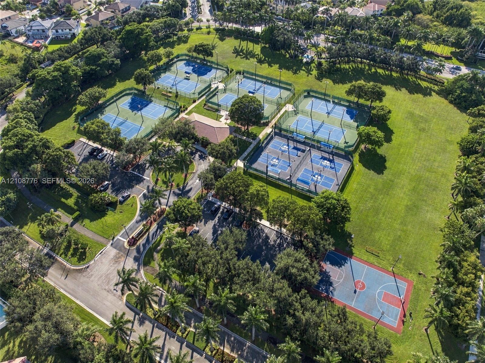 Recreational complex featuring multiple tennis courts, a paved basketball court, asphalt parking areas, and mature trees situated on expansive lawn grounds