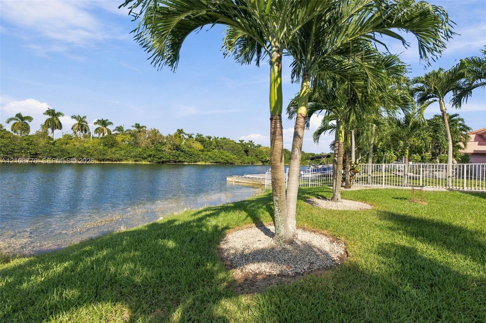 Waterfront grounds featuring a grass lawn, mature palm trees, decorative stone tree rings, and a perimeter fence