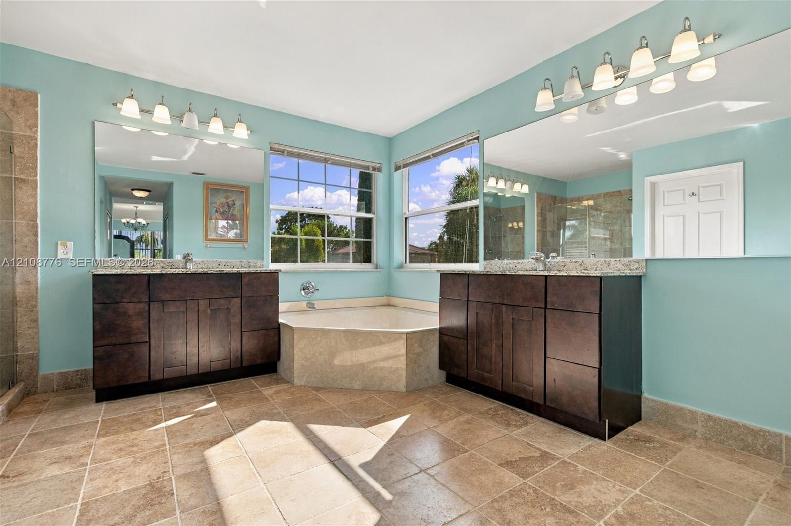 Dual vanities with dark wood-finish cabinetry, stone-surface countertops, tile flooring, corner soaking tub with stone surround, and wall-to-wall mirrors