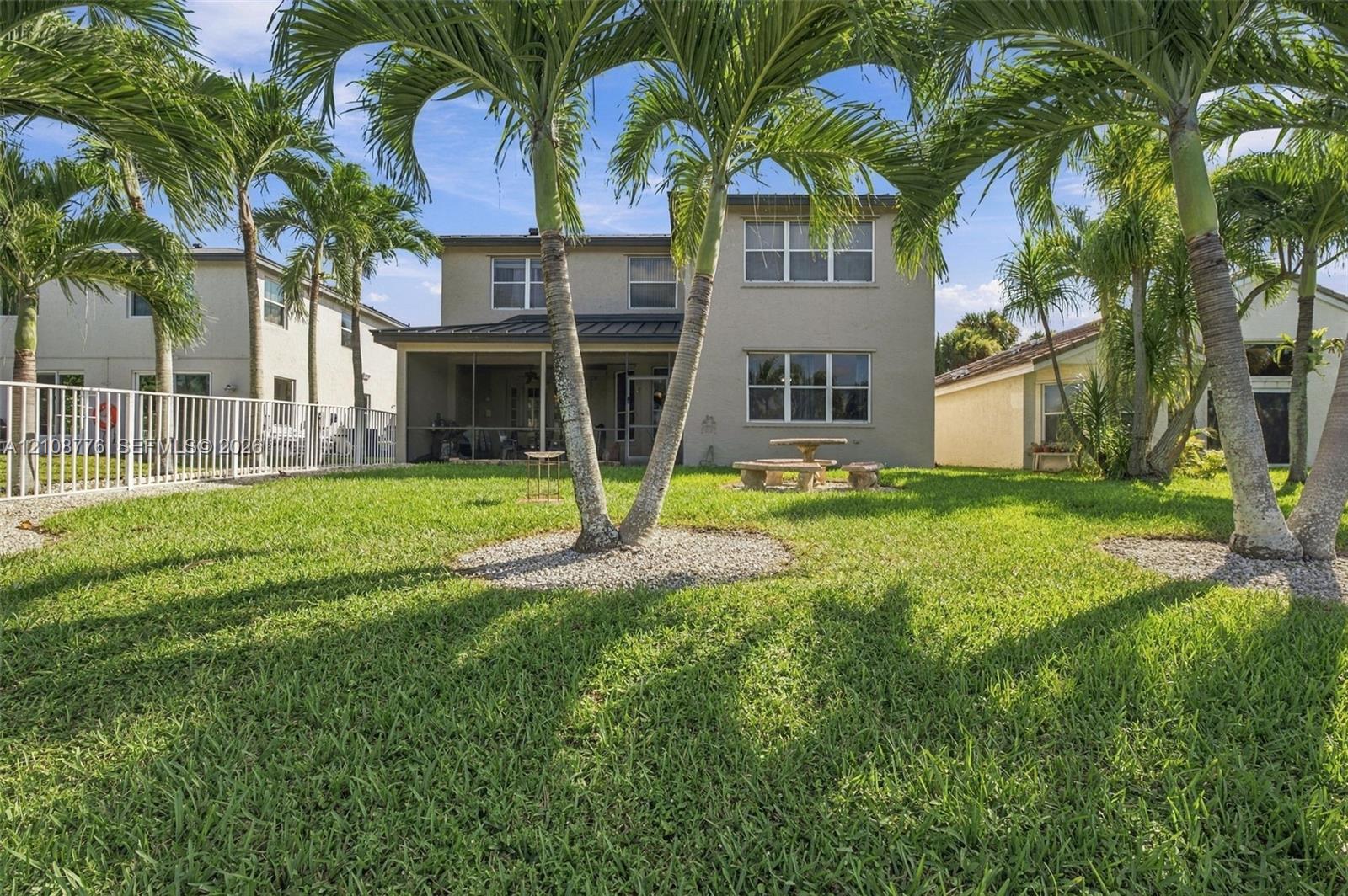 Backyard space featuring a screened-in lanai, mature palm trees, white metal perimeter fencing, and a grass lawn