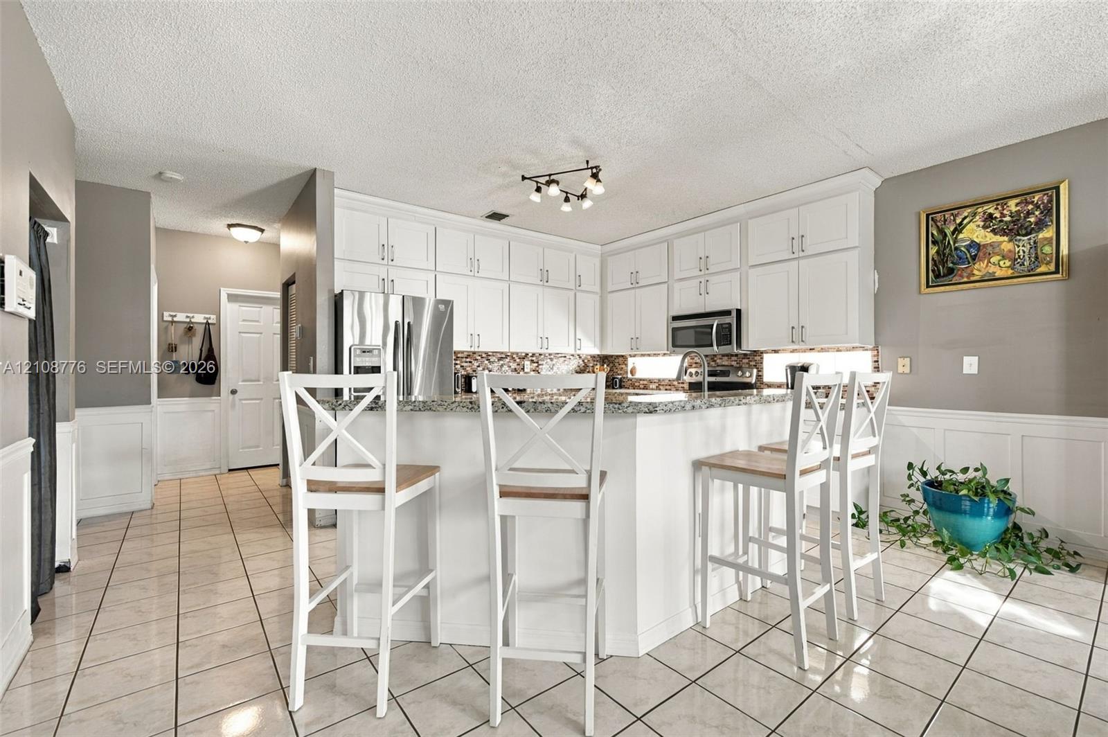 Kitchen featuring white shaker-style cabinetry, a stone countertop, a tile backsplash, and ceramic tile flooring with white wainscoting