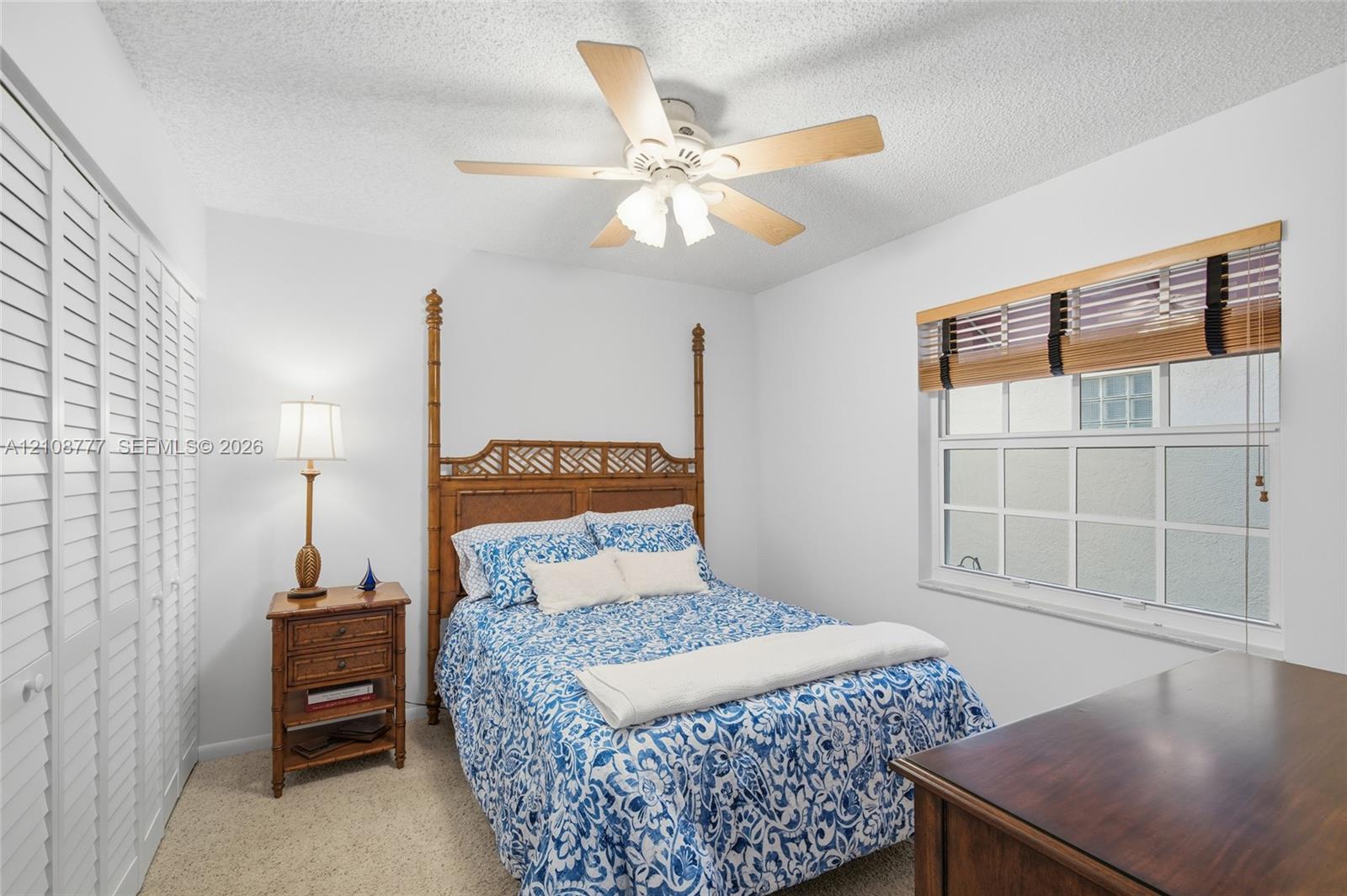 Bedroom featuring wall-to-wall louvered closet doors, multi-pane window with wood-slat blinds, textured ceiling, and a central ceiling fan with integrated light fixture