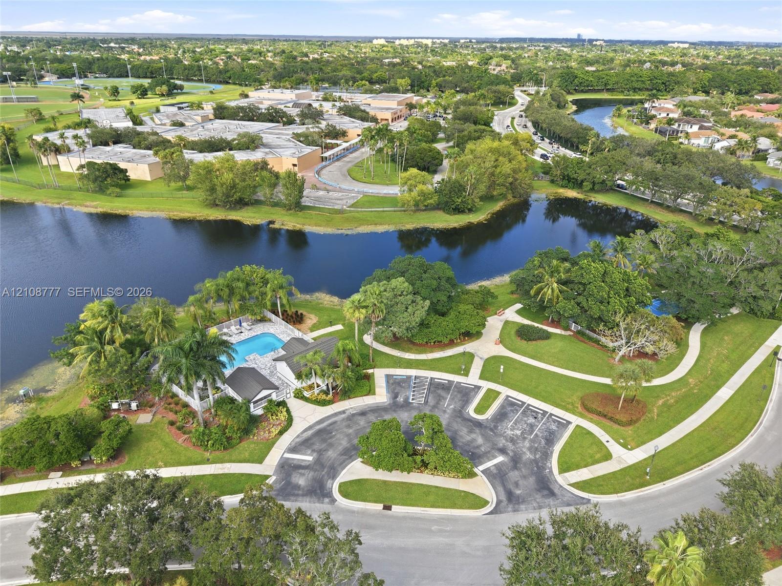 Aerial perspective featuring a waterfront swimming pool, paved parking lot, concrete walkways, and mature landscaping
