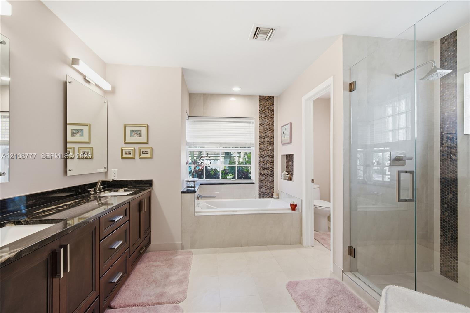 Double vanity with dark wood cabinetry and stone countertops, oversized walk-in glass shower with tile inlay, and built-in soaking tub