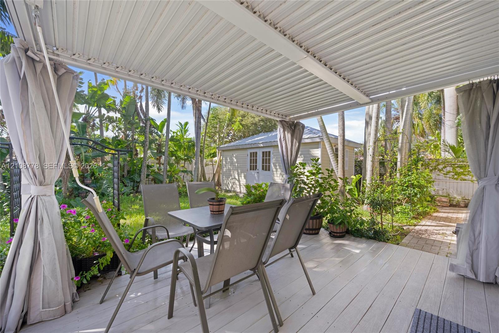 Covered deck with light-toned wood-finish flooring, metal pergola structure, and detached exterior storage building set within a mature landscape