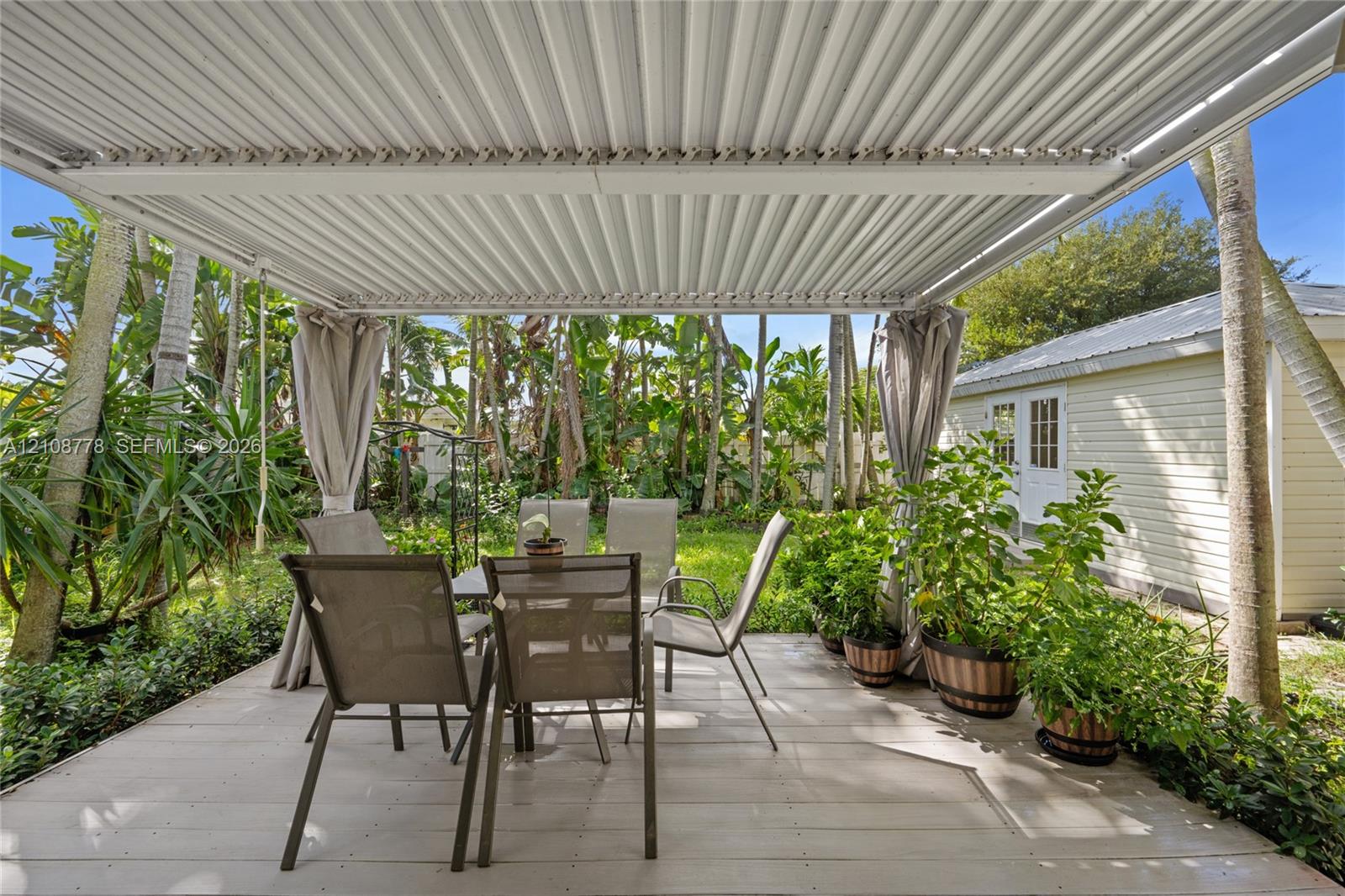 Covered patio with a corrugated metal roof structure, wood-plank style decking, and mature tropical landscaping