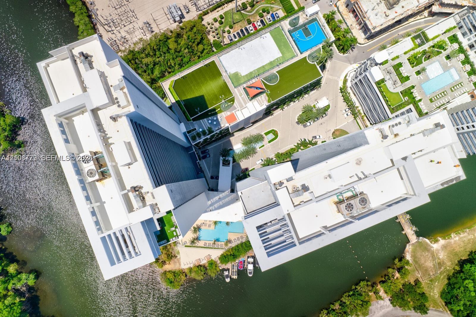 Aerial perspective of high-rise structure featuring waterfront access, outdoor swimming pool, basketball court, soccer field, and landscaped grounds