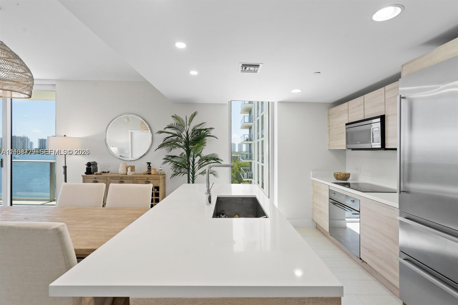 Modern kitchen featuring a central island with a solid-surface countertop and undermount sink, flat-panel light wood-finish cabinetry, stainless steel appliances, recessed ceiling lighting, and large-format tile flooring Modern kitchen featuring a central island with a solid-surface countertop and undermount sink, flat-panel light wood-finish cabinetry, stainless steel appliances, recessed ceiling lighting, and large-format tile flooring