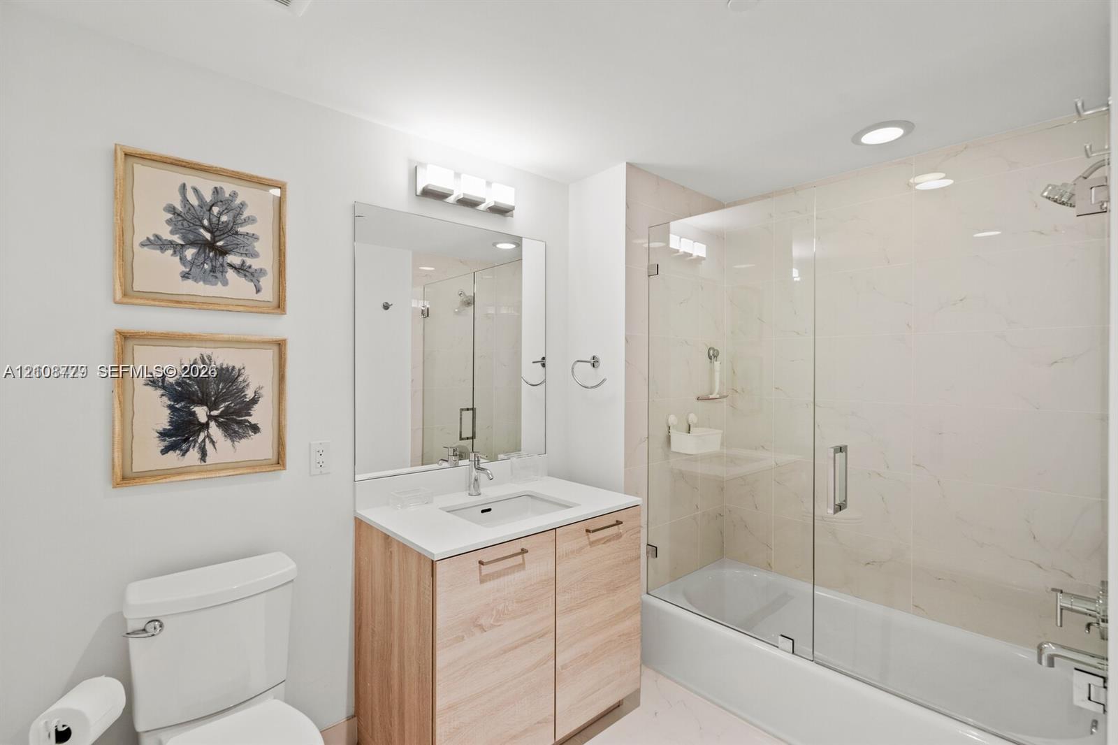 Bathroom featuring a wood-finish vanity with a rectangular sink, stone-surface countertop, full-height wall mirror, and a tub-shower combination enclosed by a frameless glass door with light-toned wall tile