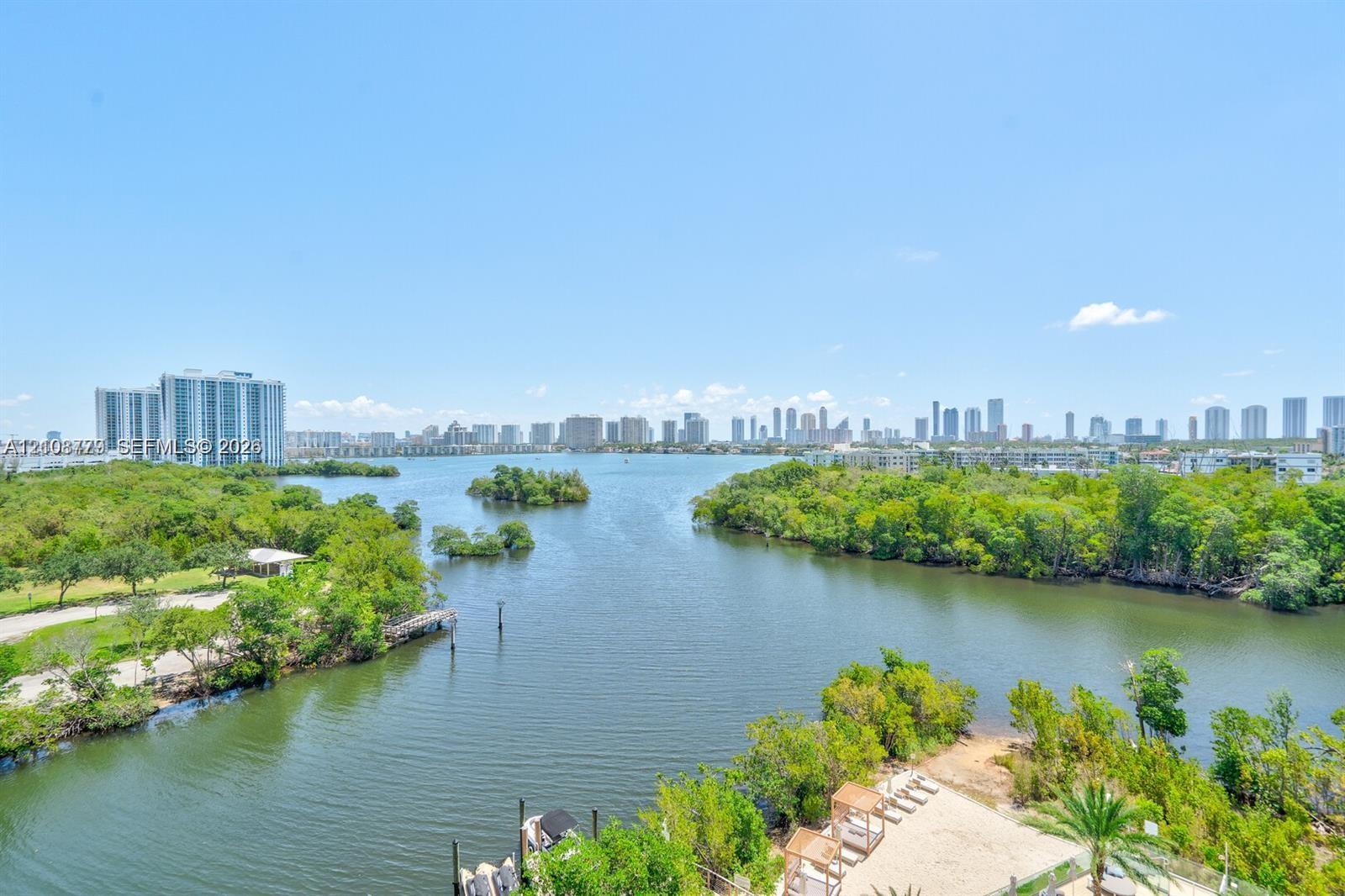 Waterfront locale featuring mangrove-lined shorelines, a sandy recreation area with shaded structures, and a distant cityscape skyline