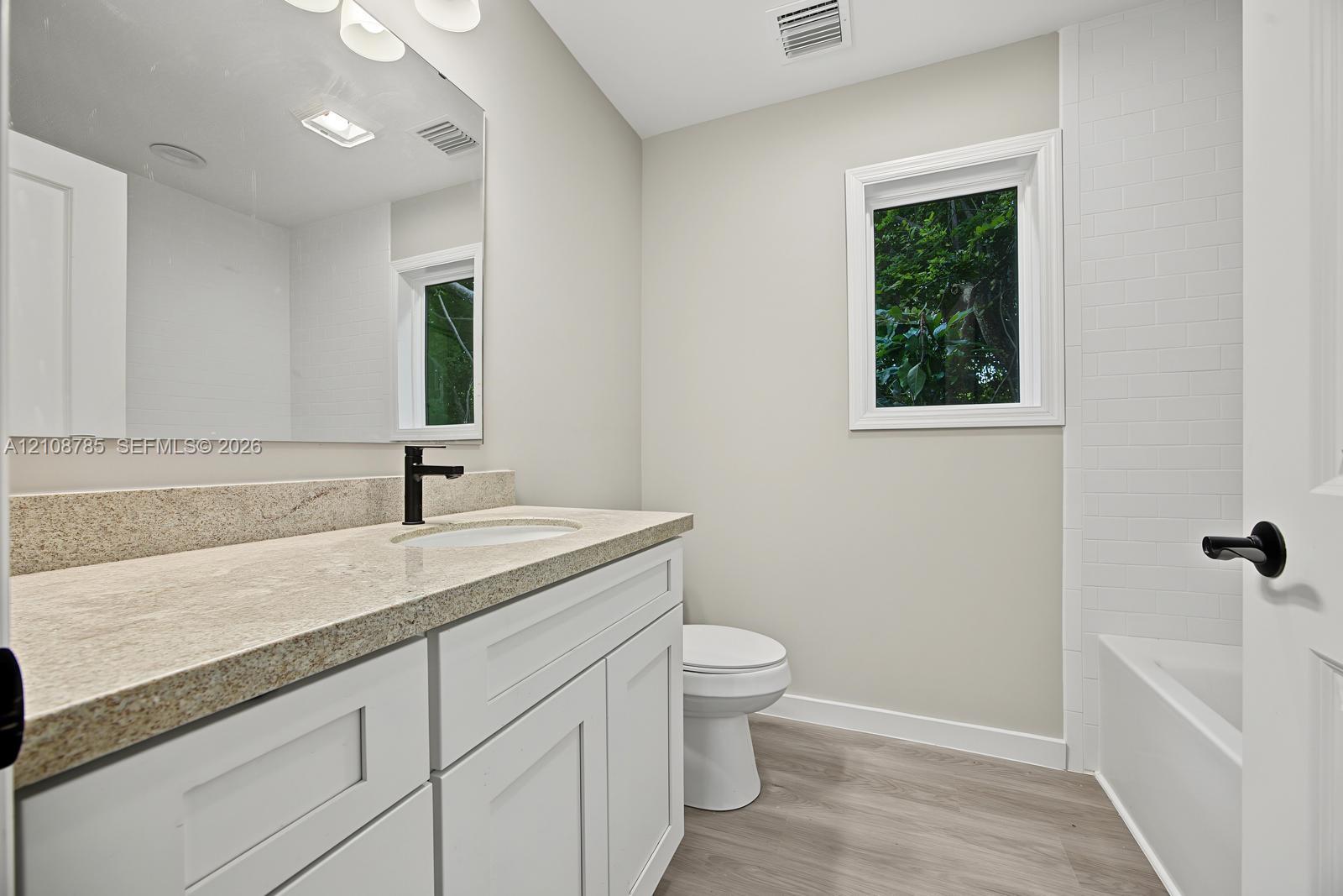Bathroom featuring a white shaker-style vanity, stone-surface countertop, dark metal hardware, wood-finish flooring, and a subway-tile tub surround