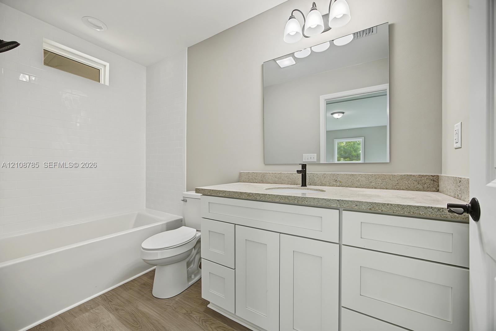 Bathroom featuring a vanity with white shaker-style cabinetry, neutral stone-finish countertop, undermount sink with dark metal hardware, and wood-finish flooring, complemented by a tiled shower surround