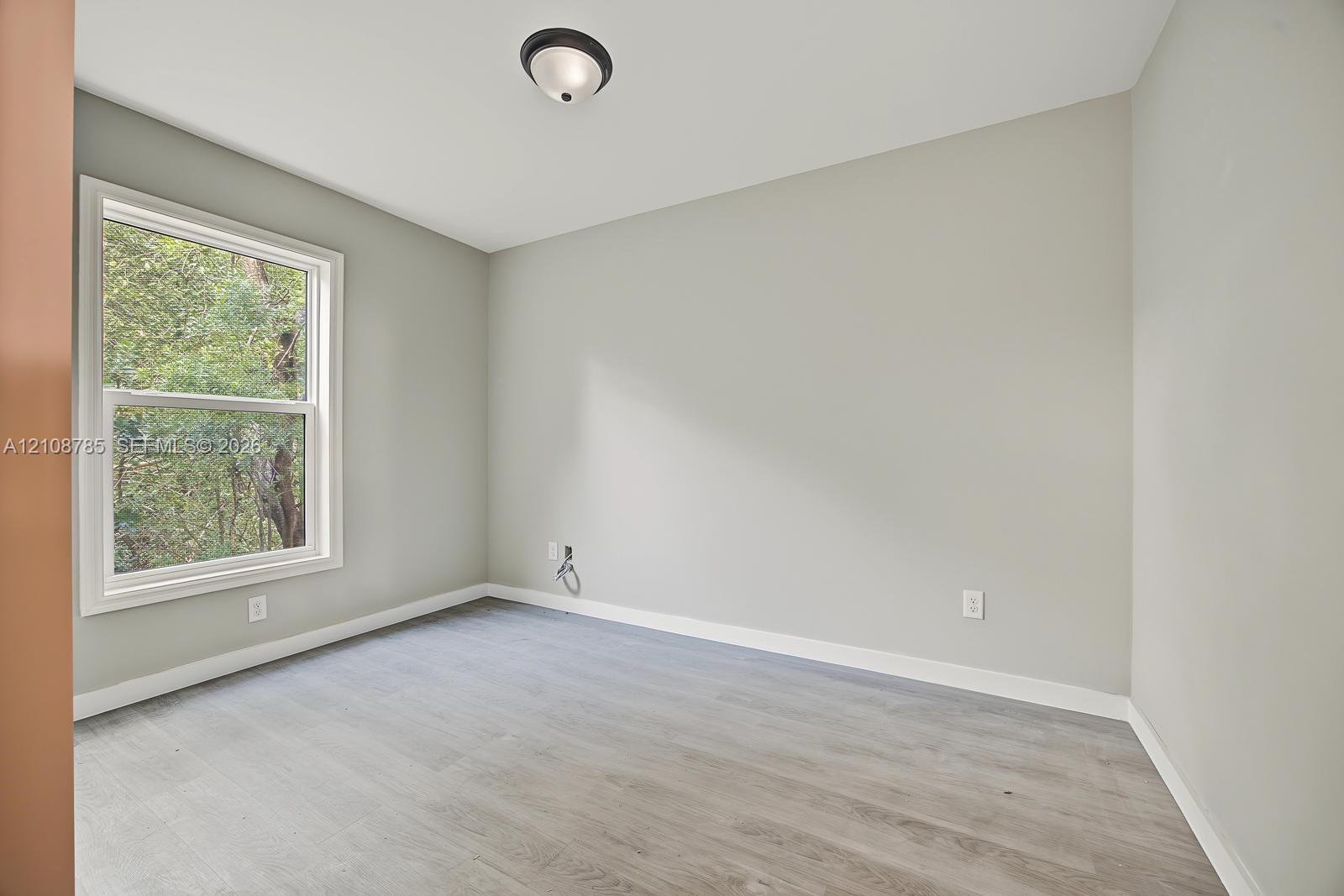 Neutral-toned interior space featuring wood-finish flooring, a single-hung window with exterior greenery, and a flush-mount ceiling light fixture