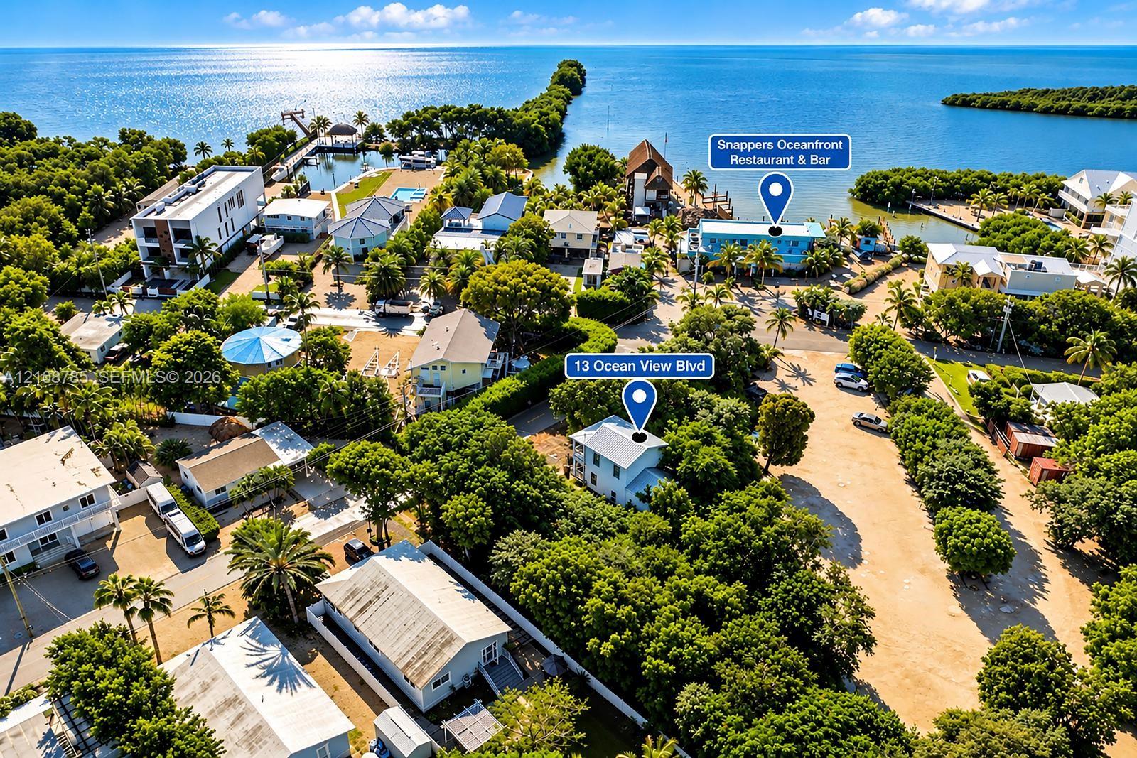Aerial perspective of coastal residential property featuring mature landscaping, metal roofing, ground-level paved parking, and proximity to the open water
