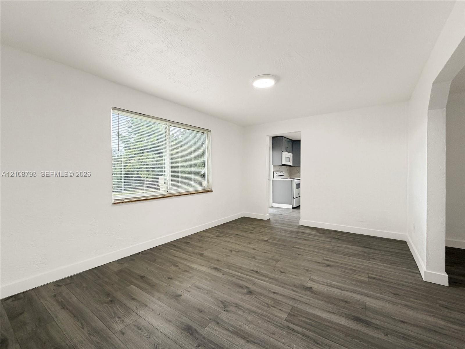 Neutral-toned interior featuring wood-finish flooring, white walls, flush-mount ceiling lighting, a large window with horizontal blinds, and a wide arched opening