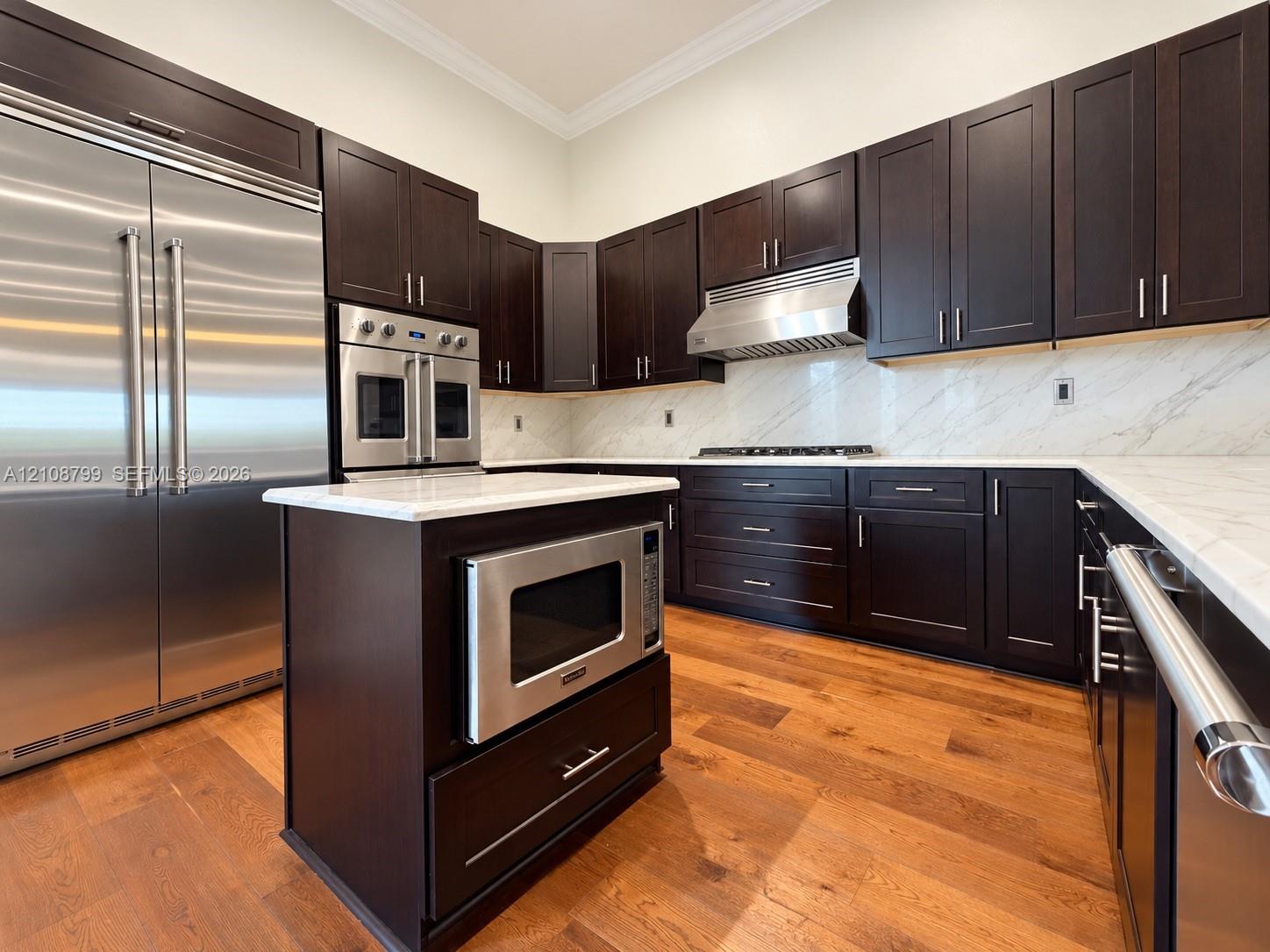 Kitchen featuring dark-finish cabinetry, stone-style countertops, stainless steel appliances, a center island with built-in microwave, and wood-finish flooring
