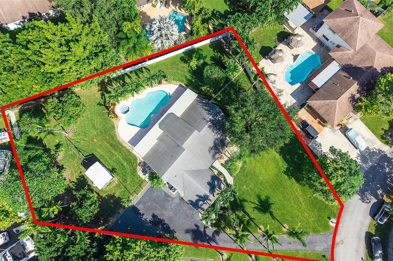 Aerial perspective of a sprawling corner lot featuring a swimming pool, expansive grass lawn, mature trees, and a paved driveway