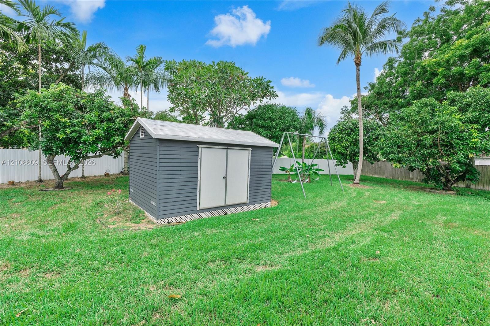 Rear yard featuring a dark gray storage shed with a gabled roof, white vinyl perimeter fencing, and mature landscaping