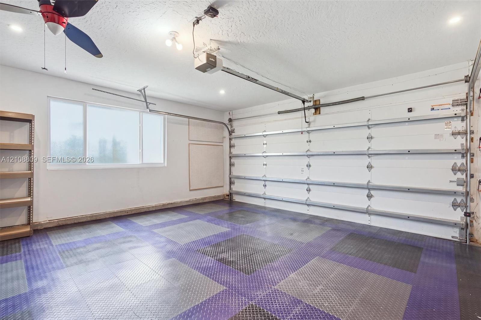 Garage space featuring patterned interlocking tile flooring, high-gloss white walls, a multi-pane horizontal window, and a ceiling-mounted fan