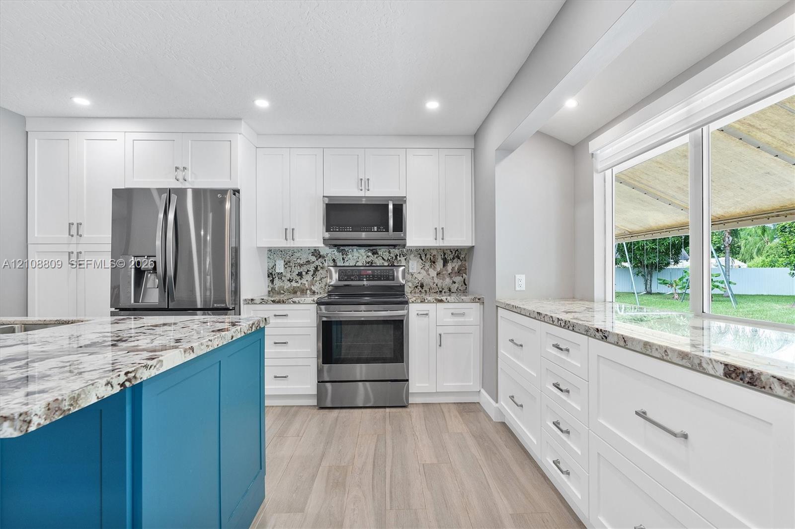 Kitchen featuring white shaker-style cabinetry, a contrasting blue island, patterned stone countertops, stainless steel appliances, and wood-finish flooring
