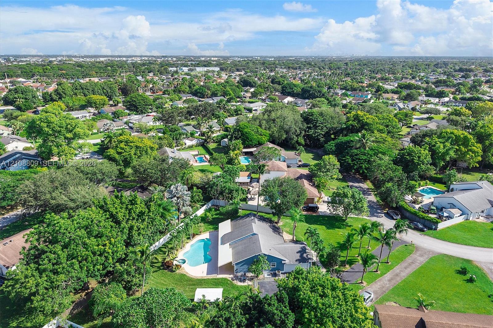Aerial perspective of residential property featuring a swimming pool, paved patio, expansive lawn, and mature trees
