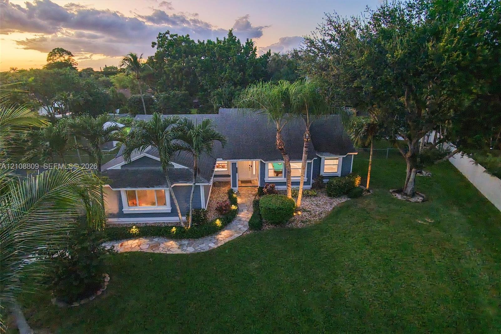 Single-story residence featuring a shingled roof, stone-paved walkway, and expansive lawn with mature trees and palms