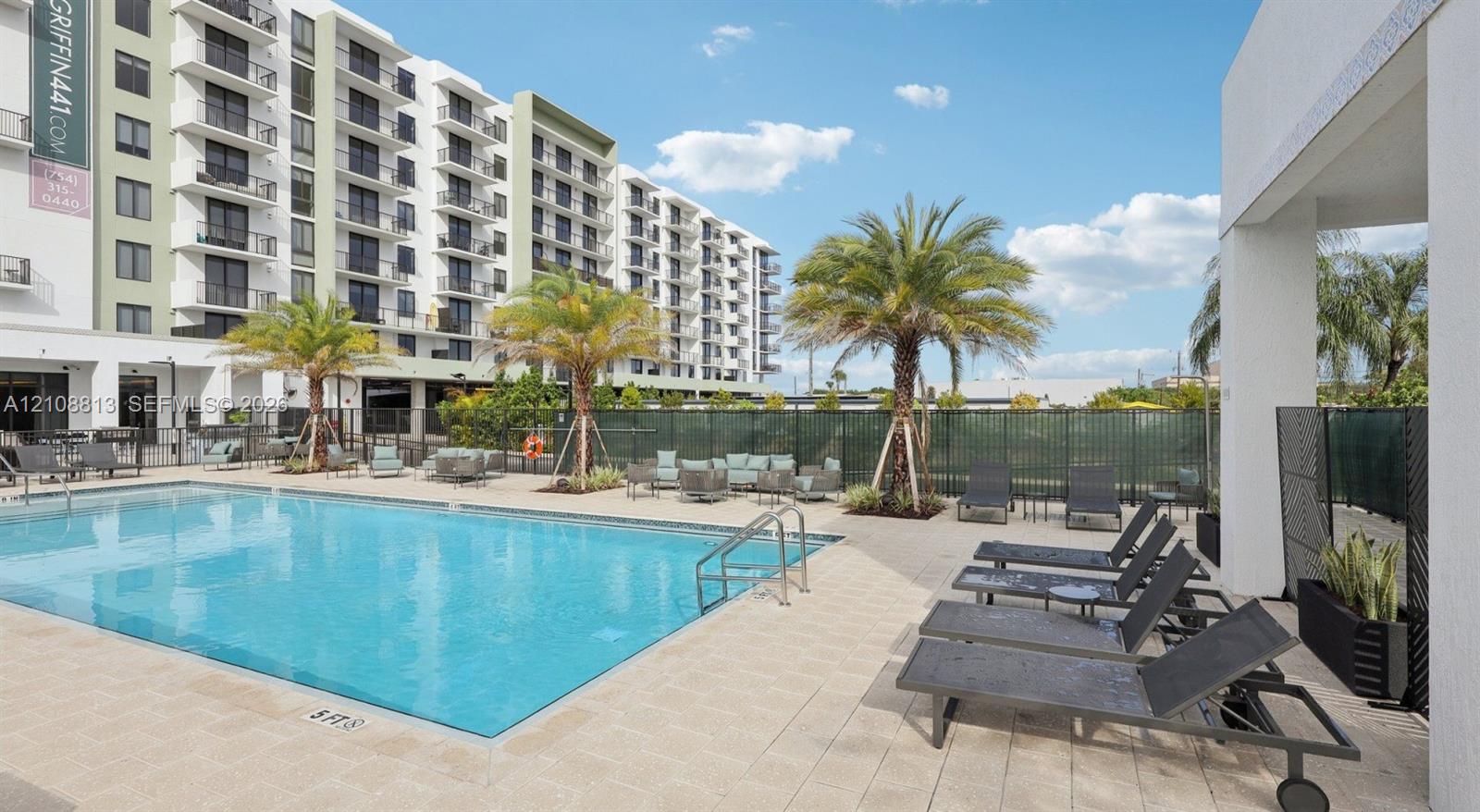 Outdoor swimming pool featuring a paved deck, surrounding palm trees, metal perimeter fencing, and concrete construction
