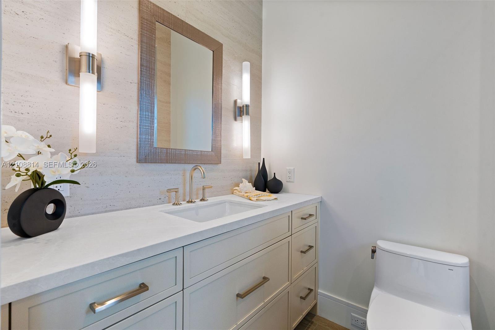 Custom vanity with quartz countertop, undermount sink, stone-finish wall cladding, dual vertical wall-mounted light fixtures, and framed mirror