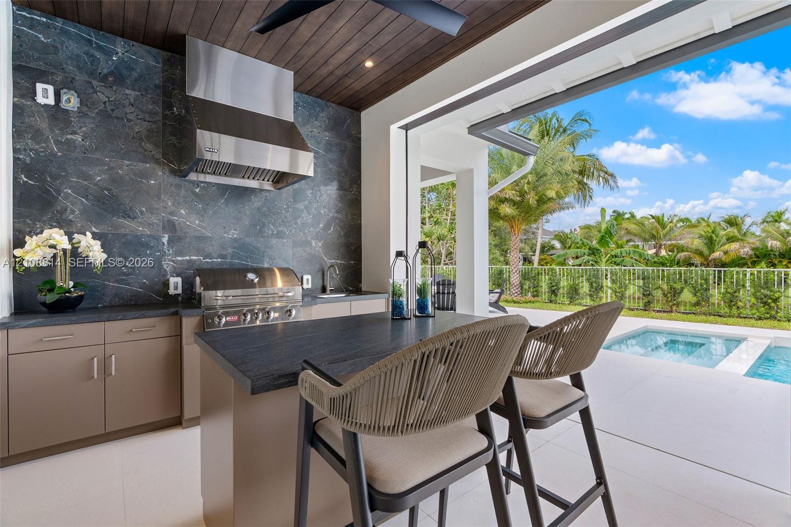 Covered outdoor kitchen featuring a dark stone backsplash, stainless steel range hood, integrated gas grill, flat-panel cabinetry, and dark-toned stone-finish countertop