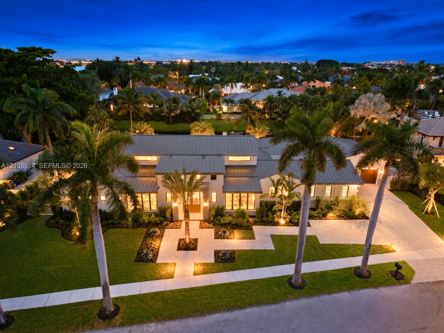 Modern residence featuring a standing-seam metal roof, paver driveway, manicured lawn, stone-surface entryway, and mature palm landscaping