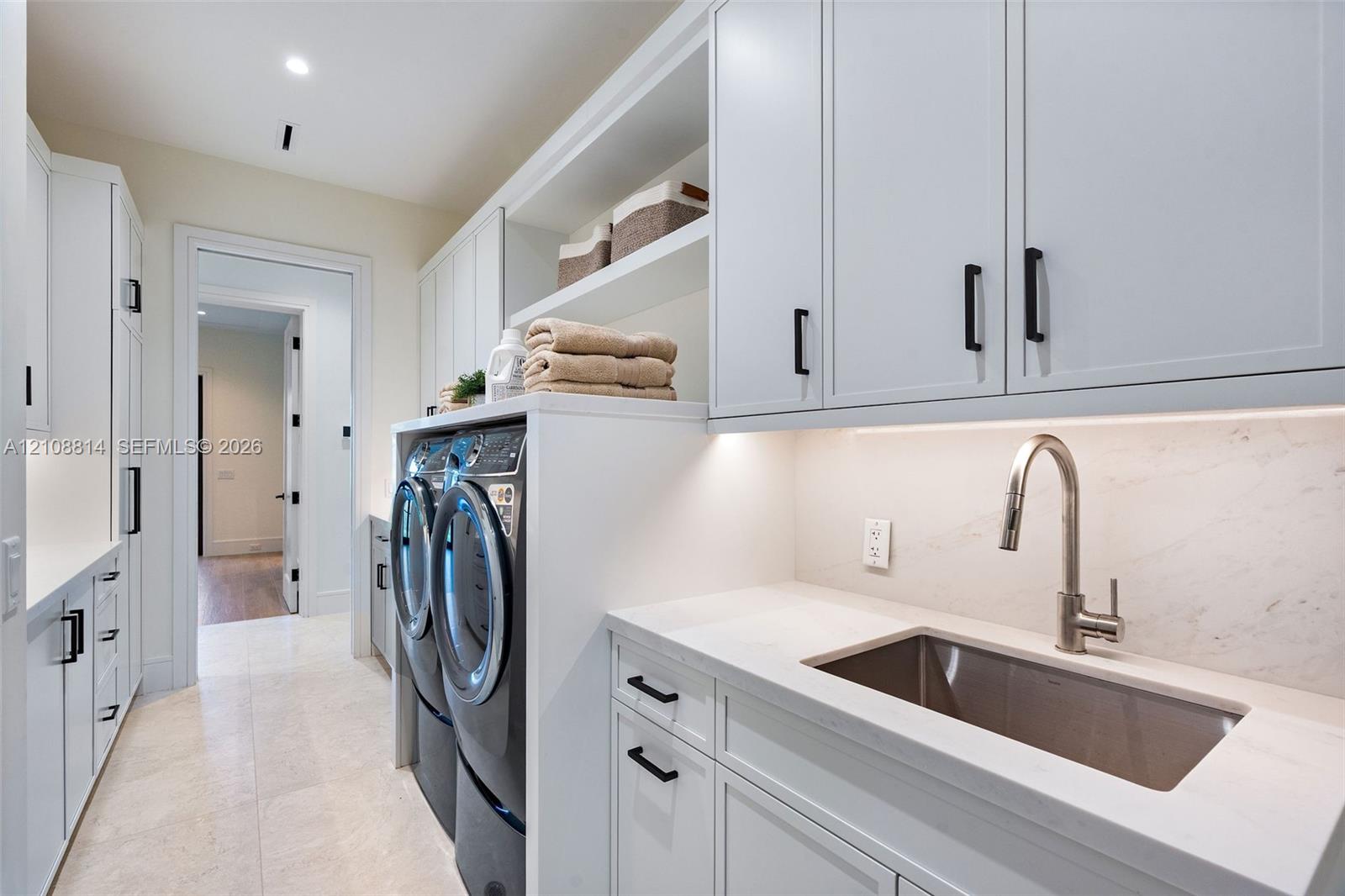 Laundry area featuring Shaker-style cabinetry, quartz countertops, a stainless steel undermount sink with a gooseneck faucet, light-toned stone-finish flooring, and recessed under-cabinet lighting