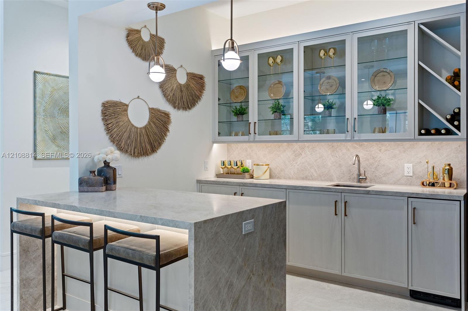 Custom cabinetry with glass-front display doors and wine storage, stone-topped island with waterfall edge, herringbone-patterned tile backsplash, undermount sink, and dual metallic pendant lighting
