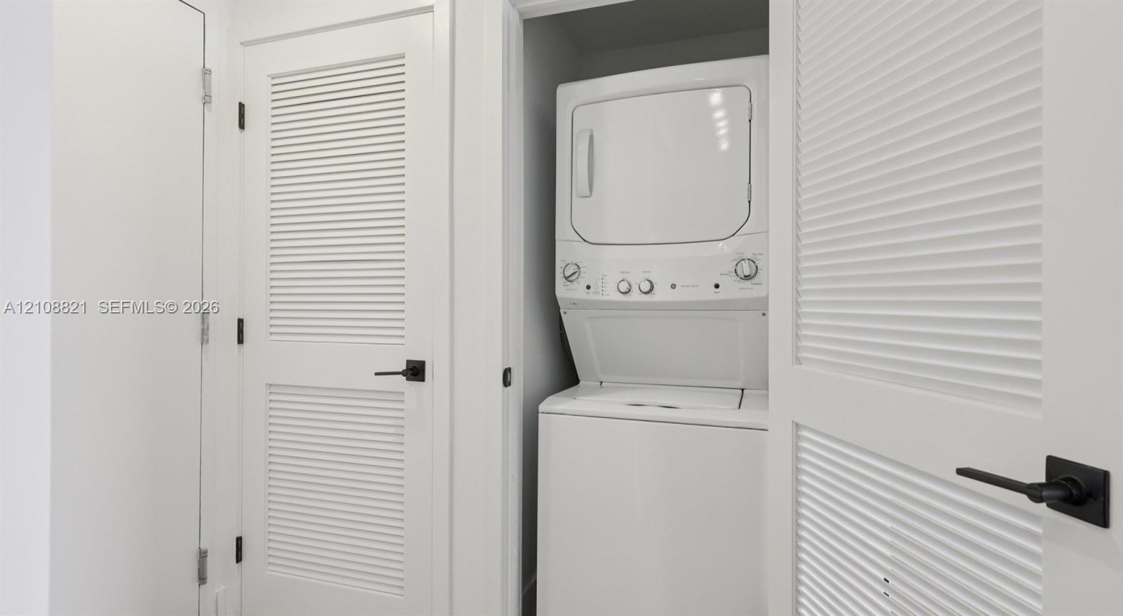 Laundry closet featuring a stackable washer and dryer unit, white louvered doors, and matte black hardware