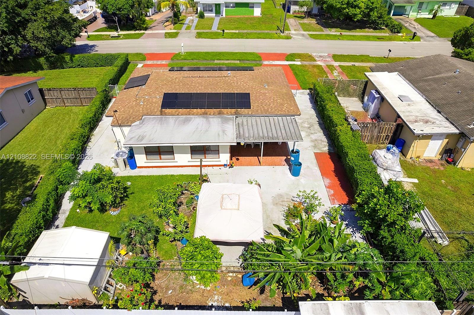 Exterior featuring a shingle roof equipped with solar panels, a covered patio, a concrete rear yard, and mature perimeter hedging