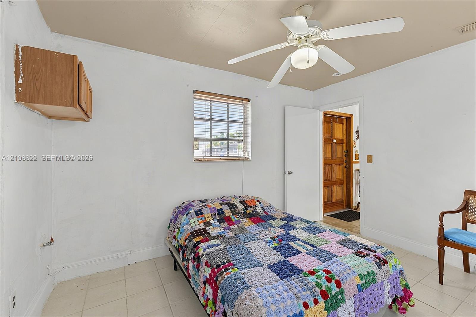 Interior room featuring light-colored tile flooring, a centered ceiling fan with integrated lighting, a wall-mounted wooden cabinet, and a single-pane window with horizontal blinds
