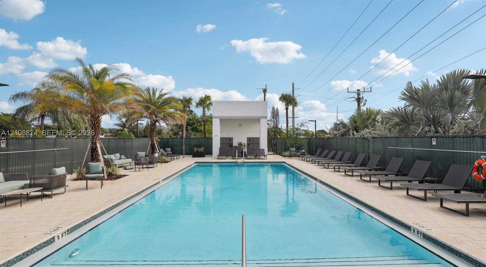Rectangular swimming pool featuring a tiled deck, perimeter fencing, palm tree landscaping, and a central cabana structure Rectangular swimming pool featuring a tiled deck, perimeter fencing, palm tree landscaping, and a central cabana structure