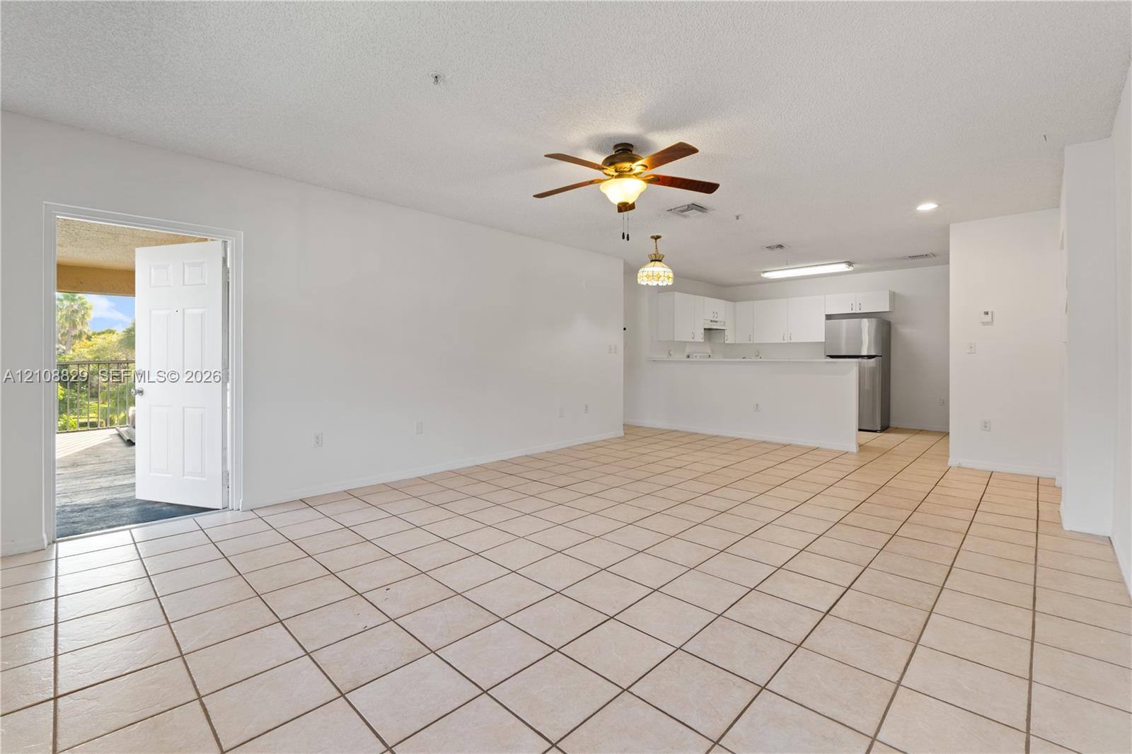 Open-concept interior featuring light-toned floor tile, a ceiling fan, white kitchen cabinetry, and a breakfast bar