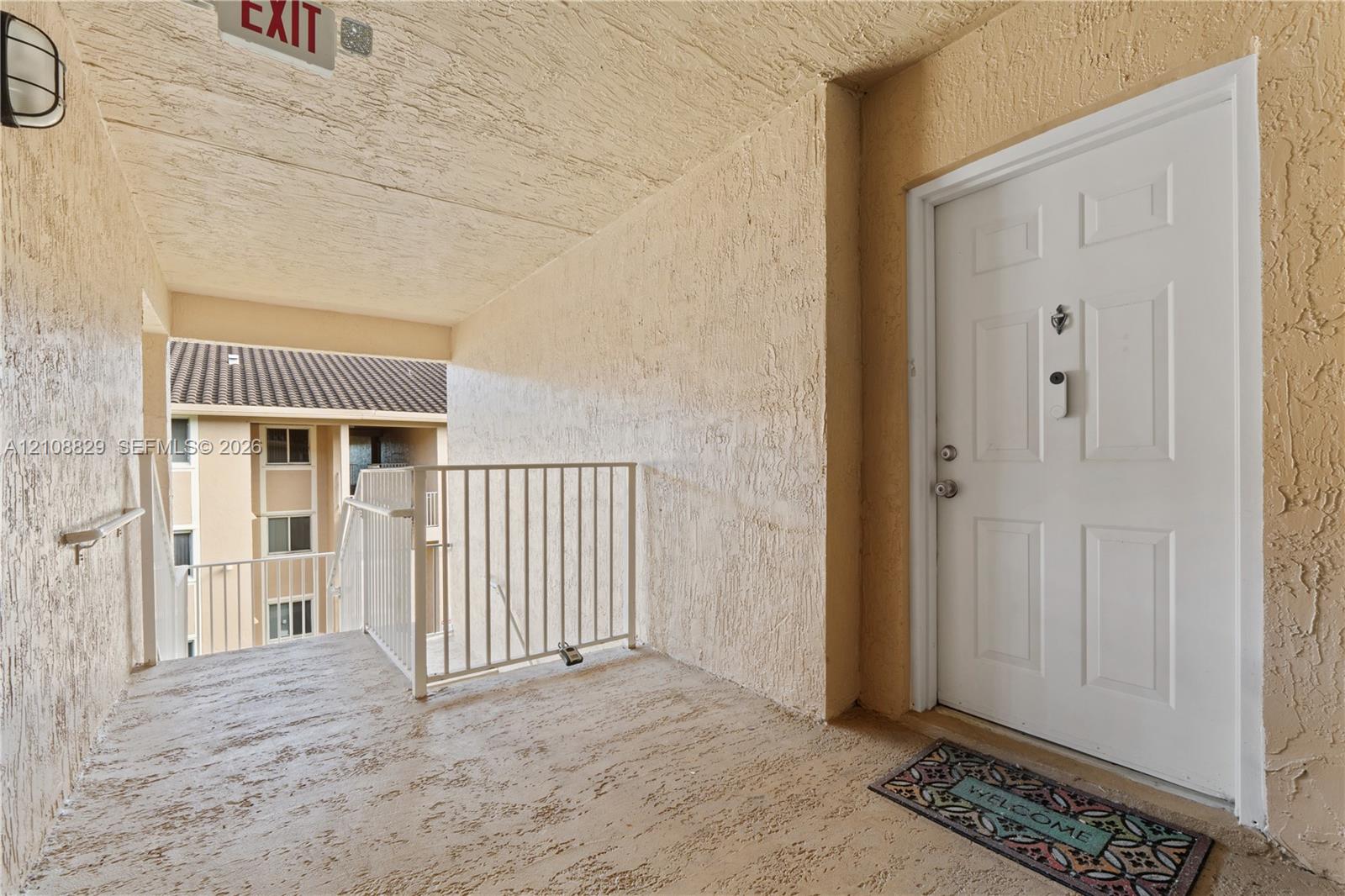 Covered landing area featuring textured stucco walls, concrete flooring, white metal railing, and a paneled entry door with hardware
