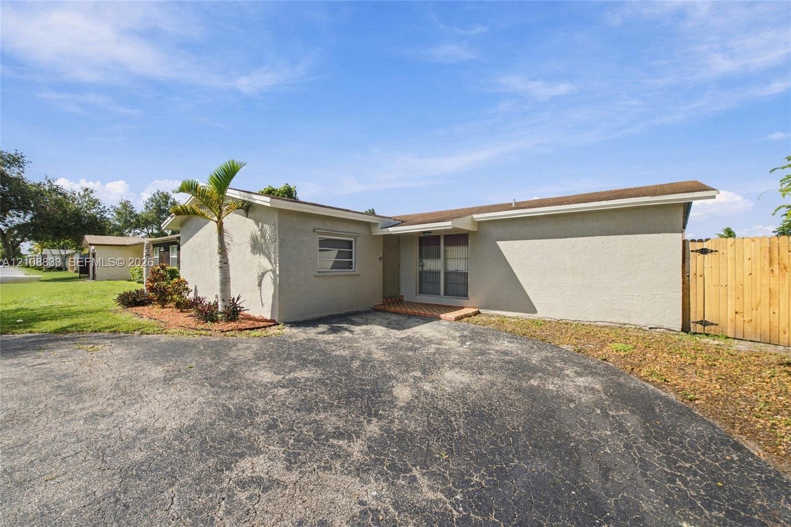 Single-story exterior featuring a light-toned stucco facade, expansive asphalt driveway, and tall wood privacy fencing