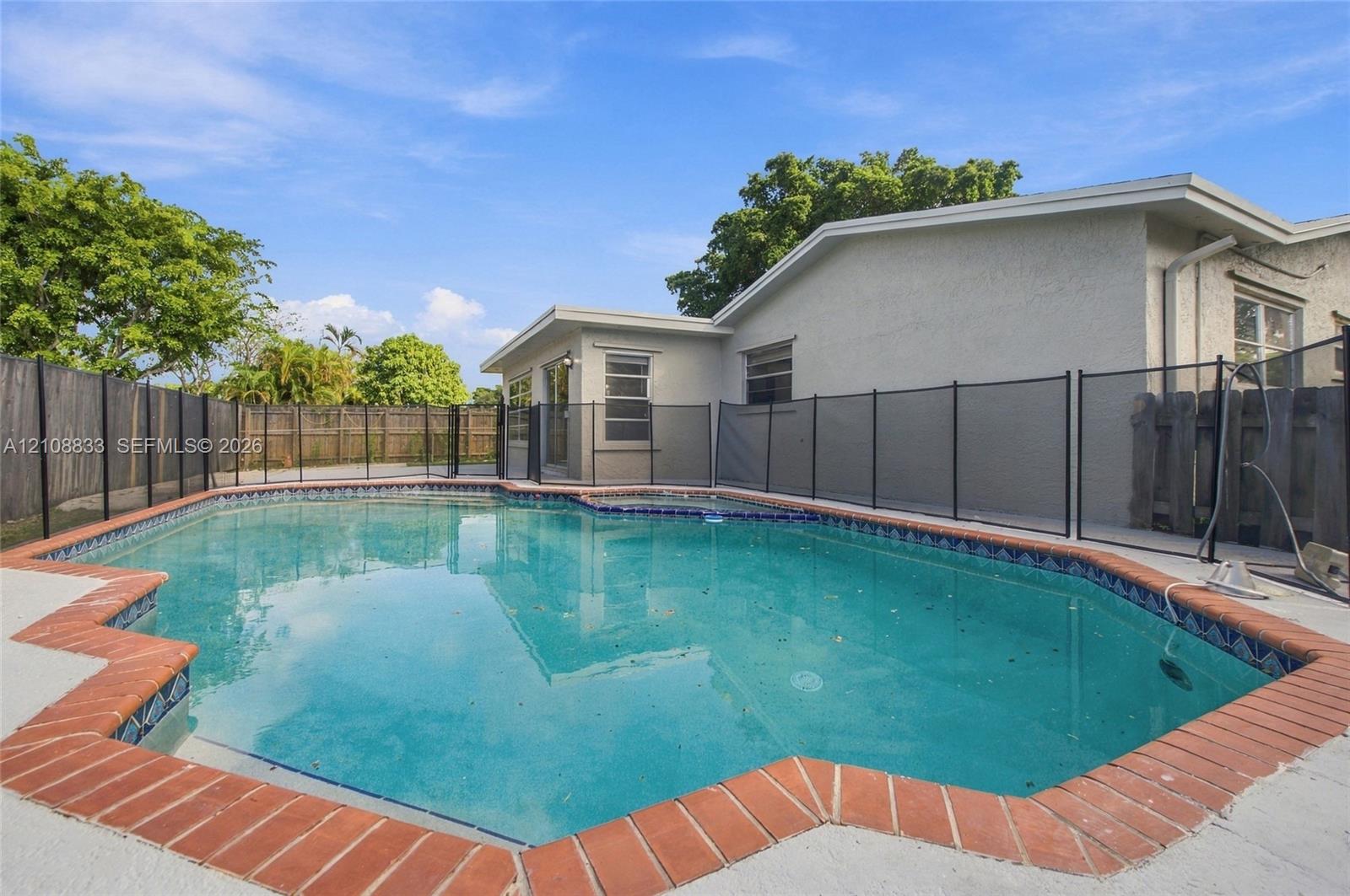 In-ground swimming pool featuring a brick paver surround, decorative tile border, and a black mesh safety fence enclosed by wood privacy fencing