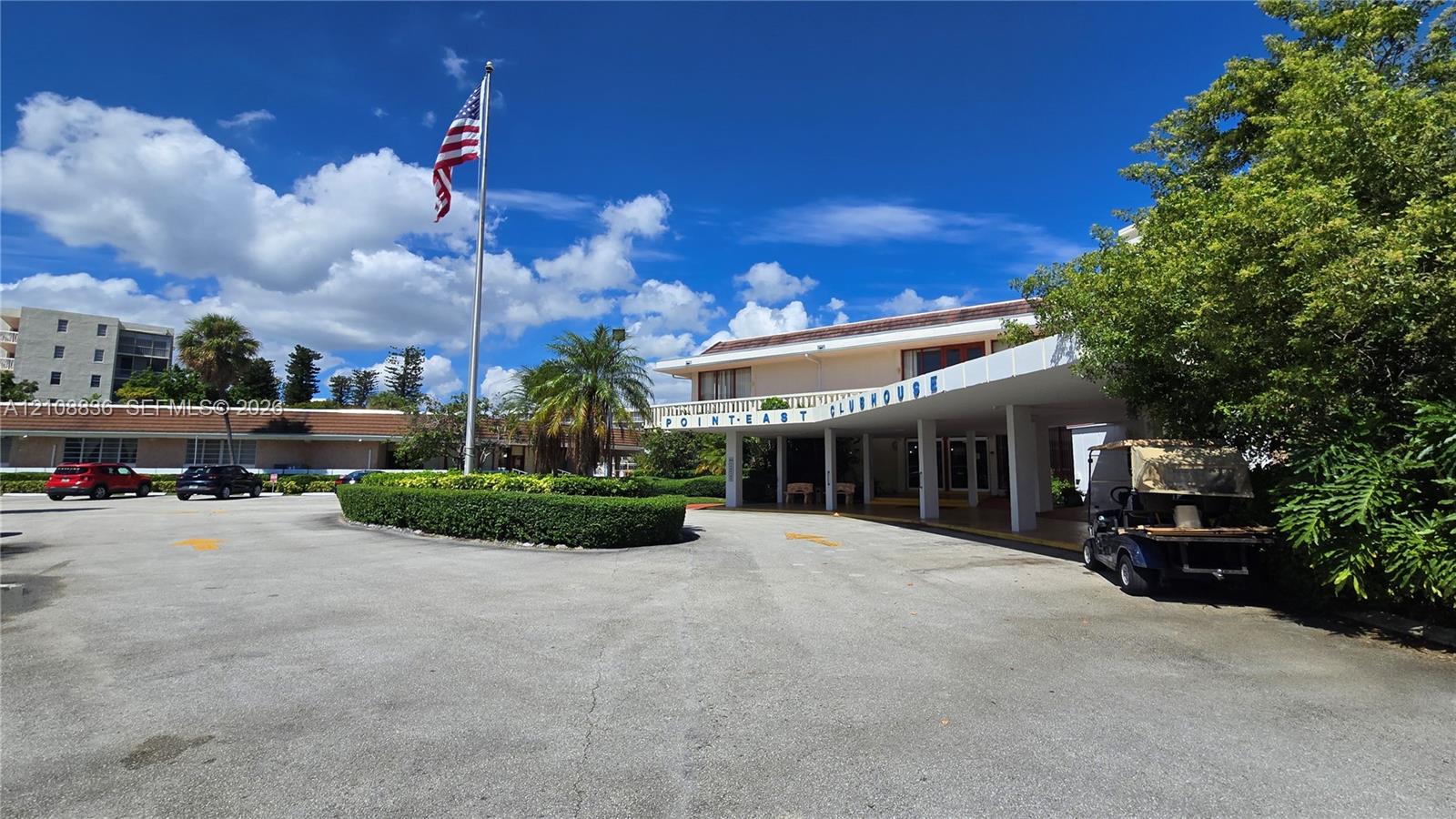 Clubhouse exterior featuring a covered porte-cochre, paved parking area, and mature landscaping