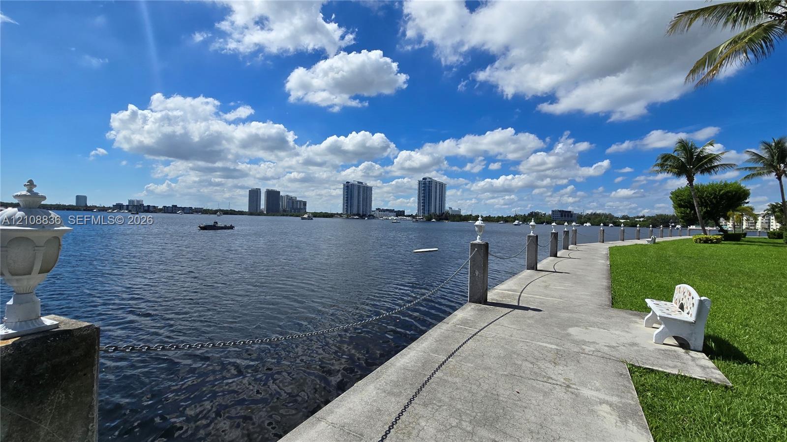 Waterfront concrete walkway featuring a post-and-chain barrier, decorative pedestal lighting, and manicured lawn